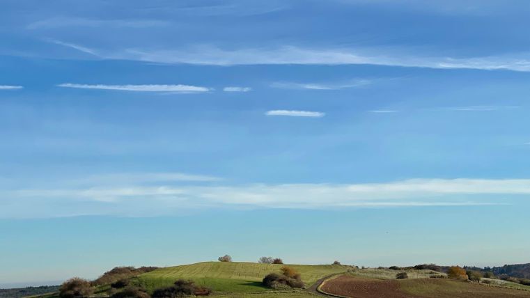 A gentle hilly landscape under a blue sky with a few clouds. In the foreground, there are green meadows and a fenced field.