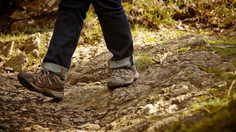 Ein Fußgänger trägt robuste Wanderschuhe und geht über einen unebenen, steinigen Weg. Um ihn herum ist die Natur mit grünem Moos und Bäumen zu sehen.