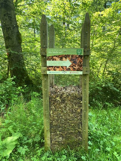 A lattice container in the forest, filled with leaves, with a sign above it.