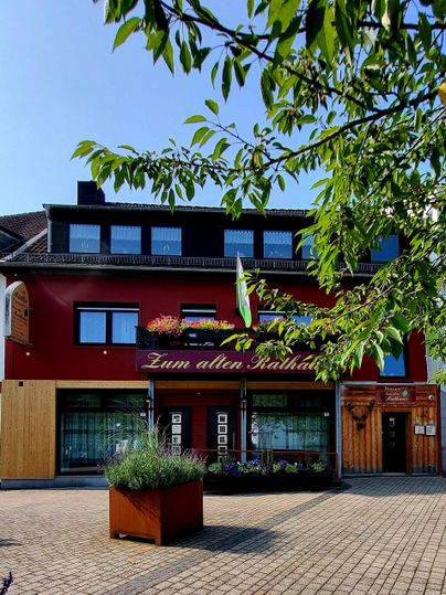 A cozy inn with a red facade and an inviting terrace. Surrounded by trees and a paved square.