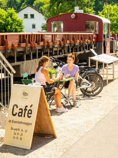 Ein Café im Eisenbahnwagen mit zwei Frauen, die an einem Tisch sitzen. Im Hintergrund sind Fahrräder und eine sonnige Landschaft zu sehen.