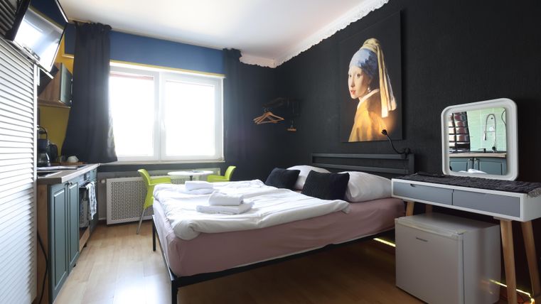 A modern room with a double bed and an artistic mural. There is a desk, a refrigerator, and plenty of light coming through the window.