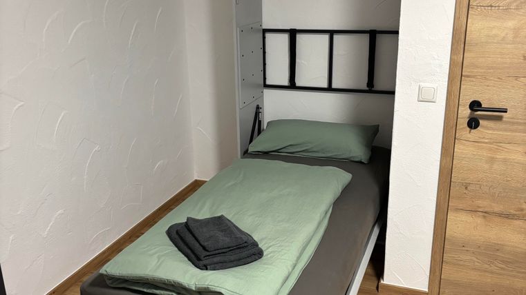 A small, modern bedroom with a foldable bed. The bed has green bedding and some towels on it.