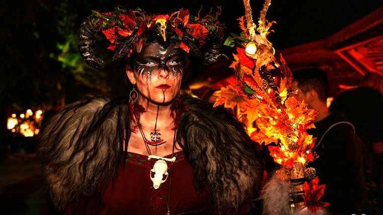 Eine Frau in auffälligem Kostüm mit einem Kopfteil aus Hörnern und bunten Blättern. Sie hält ein leuchtendes Accessoire und hat ein dramatisches Make-up.