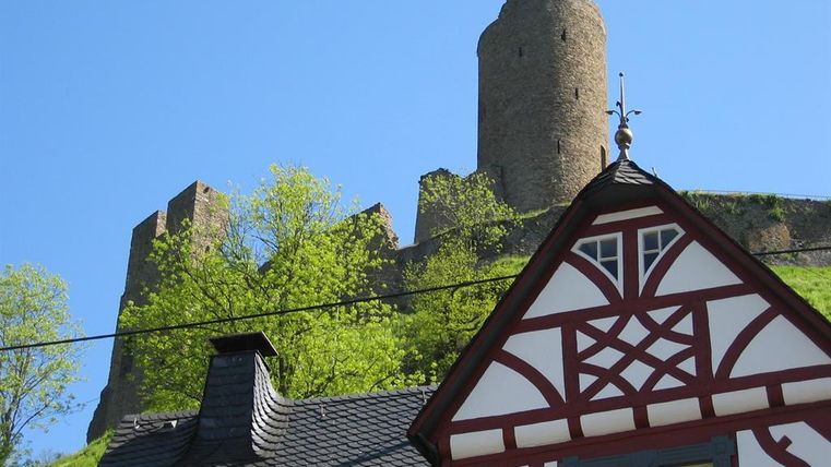 Eine alte Burg steht auf einem Hügel, umgeben von frischem Grün. Im Vordergrund ist ein traditionelles Fachwerkhaus zu sehen.