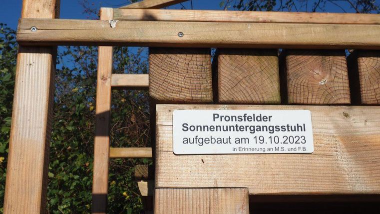 Een houten stoel met de tekst "Pronsfelder Zonnebankstoel", die op 19.10.2023 is opgezet. Op de achtergrond zijn bomen en een blauwe lucht te zien.