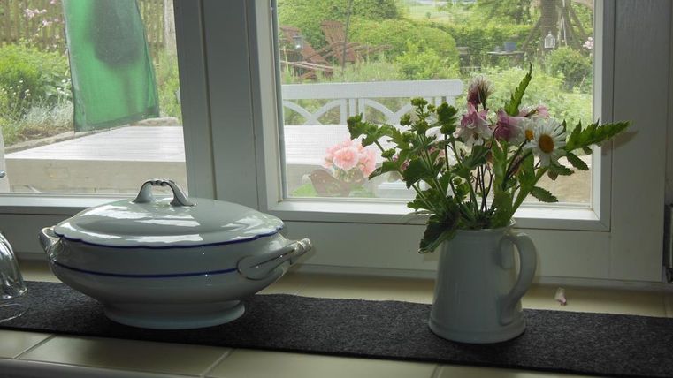 Ein schöner Fensterbereich mit einem weißen Geschirrtopf und einer Vase voller bunter Blumen. Draußen ist eine grüne Landschaft zu sehen.