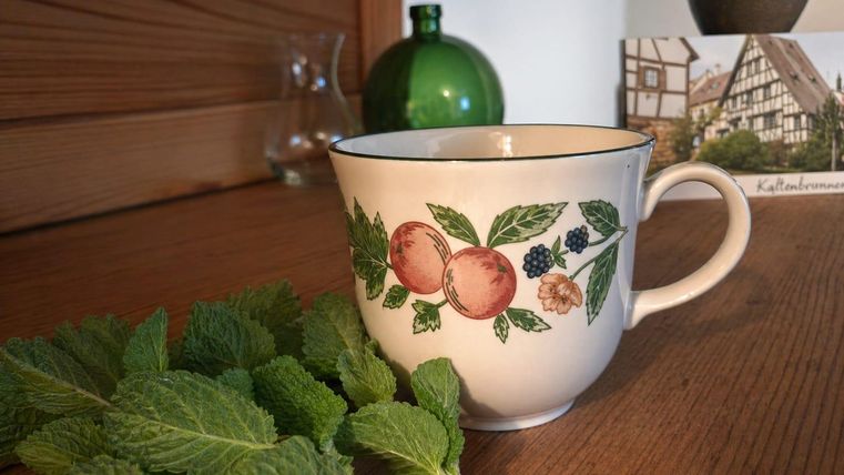 A cup with a colorful fruit pattern sits on a wooden table. Next to the cup lies a fresh mint leaf.