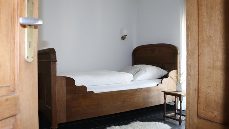 Une chambre simple avec un lit en bois et des draps blancs. À côté du lit se trouve une petite table et un tapis est posé sur le sol.