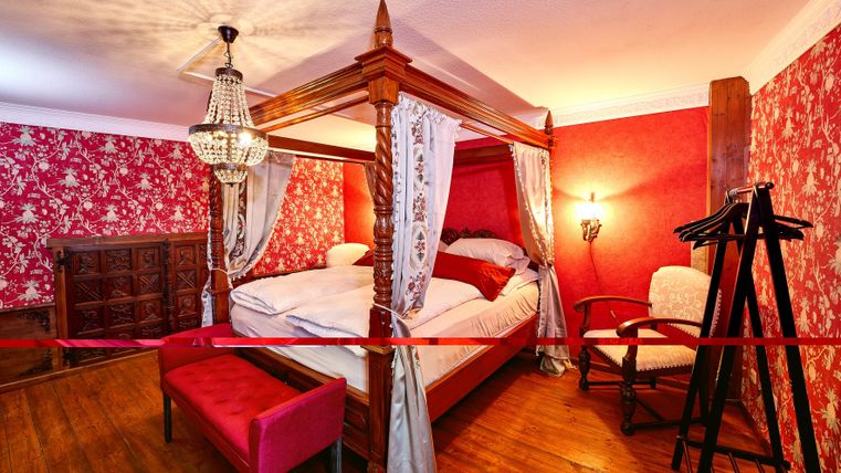 Ein elegantes Schlafzimmer mit einem Himmelbett, umgeben von einer roten Tapete mit floralem Muster. Im Raum befinden sich ein Stuhl und ein Kleiderständer.