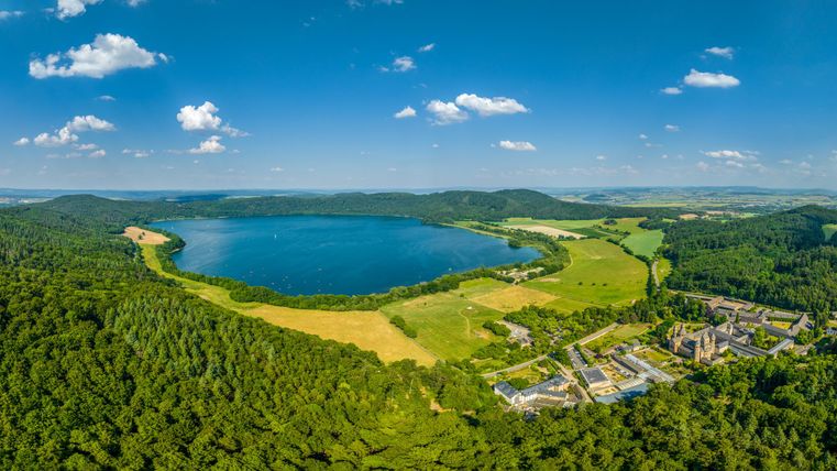 Ein weitläufiger Blick auf einen glitzernden See, umgeben von dichten Wäldern und saftigen Wiesen. Im Hintergrund sind sanfte Hügel und ein paar Wolken am blauen Himmel zu sehen.