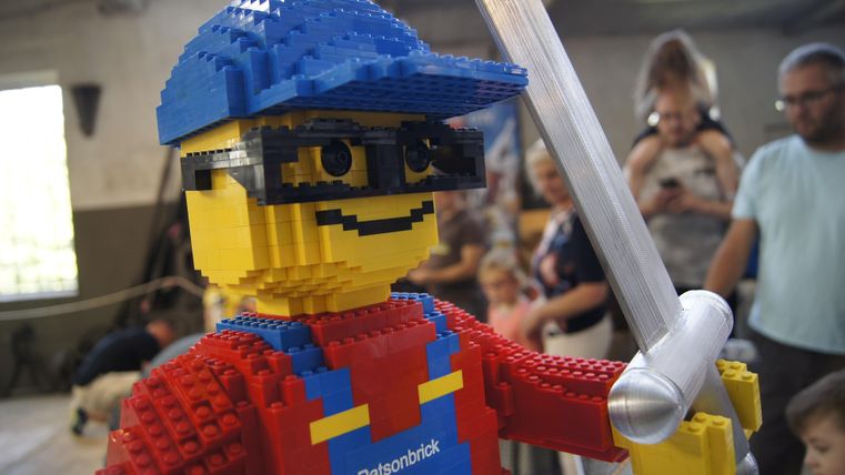 Ein großer LEGO-Charakter mit blauer Mütze und rotem Shirt hält ein Schwert in der Hand. Im Hintergrund sind Menschen zu sehen, die ihn betrachten.