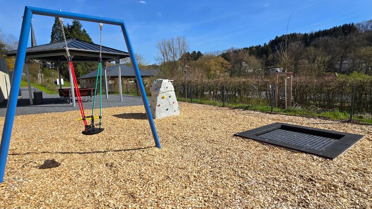A beautiful playground with a swing, a climbing wall, and a trampoline. The sky is clear and there is plenty of space to play.
