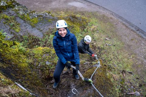Two people are climbing a steep, grassy rock face. Both are wearing helmets and are equipped with climbing gear.