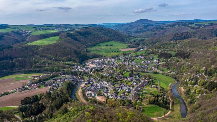 A picturesque view of a small village in a hilly landscape. Surrounded by green fields and a flowing river.