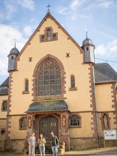 Een oude kerk met een mooie gevel en twee torens. Voor staan drie personen die ontspannen met elkaar praten.