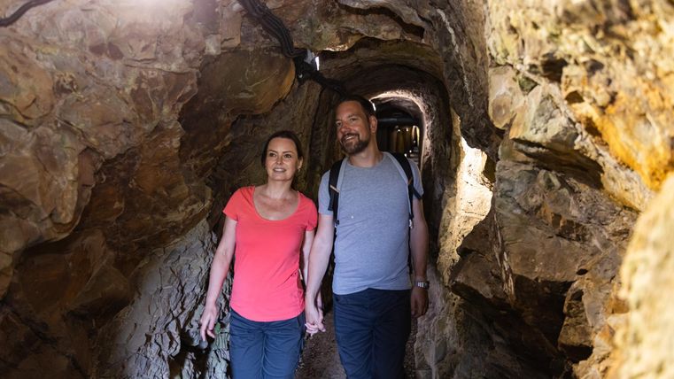 Un couple marche main dans la main à travers un étroit tunnel de pierres éclairé. L'atmosphère semble aventureuse et intéressante.