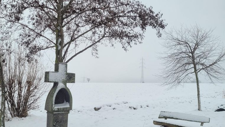 Eine verschneite Landschaft mit einem Denkmal und einem Baum. Neben dem Denkmal steht eine Bank und ein kleiner Tisch.