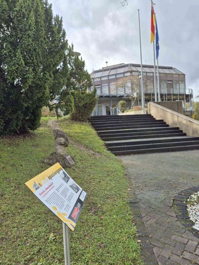 Een modern gebouw met een glazen gevel en twee vlaggen ervoor, omringd door bomen en een uithangbord op de voorgrond.