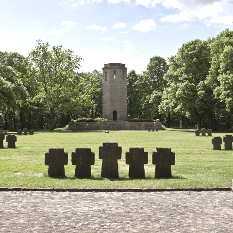 Ein Kriegsdenkmal mit Steinkreuzen auf einer Wiese, ein Turm im Hintergrund, umgeben von grünen Bäumen unter blauem Himmel., © TI Bitburger Land