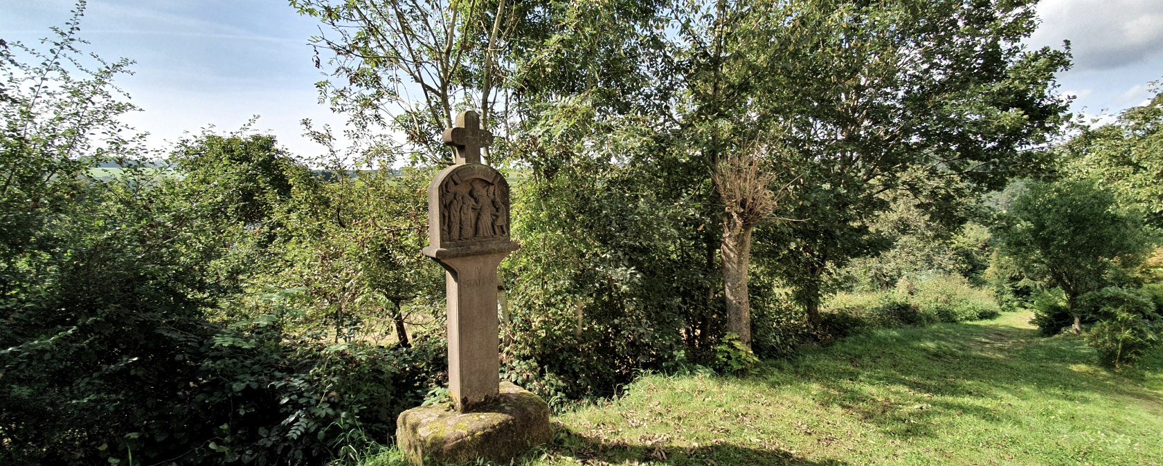 Une croix en pierre se dresse sur une pelouse verte, entourée d'arbres et d'arbustes. Le ciel est bleu avec quelques nuages., © Tourist-Information Bitburger Land