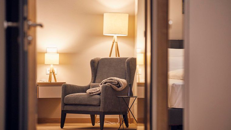 Een gezellige fauteuil staat in een moderne kamer met warm licht. Op de achtergrond is een bed met een nachtkastje te zien.