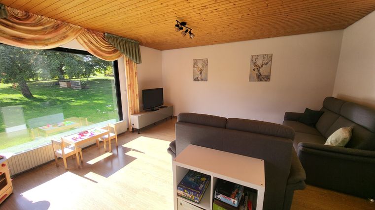 Een lichte woonkamer met een houten plafond en grote ramen. Er is een bank, een televisie en een kleine tafel voor kinderen.