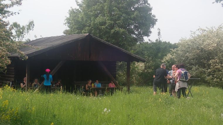 Eine kleine Gruppe von Menschen versammelt sich vor einer Holzunterkunft in einer grünen Wiese. Im Hintergrund sind Bäume und blühende Sträucher sichtbar.