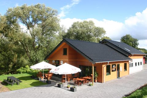 Houten gebouw met terras, parasols en zitgelegenheden, omringd door bomen en een blauwe lucht.