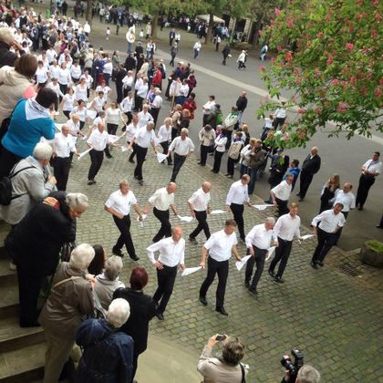 Eine Gruppe von Männern in weißen Hemden tanzt synchron auf einem Platz. Im Hintergrund stehen viele Zuschauer und beobachten die Darbietung.