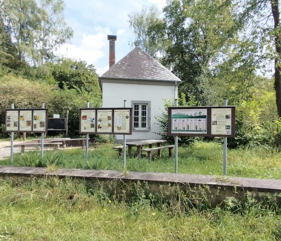 Een klein wit gebouw met informatieborden en bankjes ervoor, omringd door bomen en gras., © TI Bitburger Land