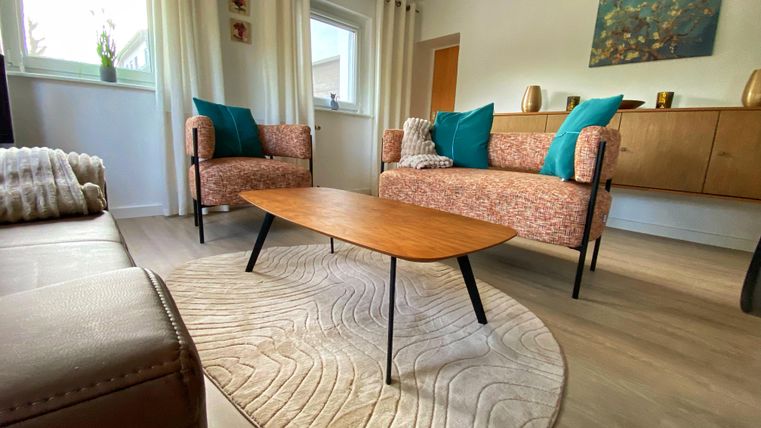 Un salon chaleureux avec deux fauteuils confortables et une table basse en bois. Les murs sont peints en clair, et il y a de grandes fenêtres avec des rideaux.