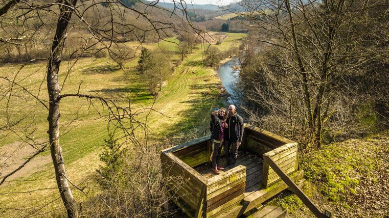 Deux personnes prennent un selfie sur un point de vue avec vue sur la boucle de la Prüm et le paysage environnant.