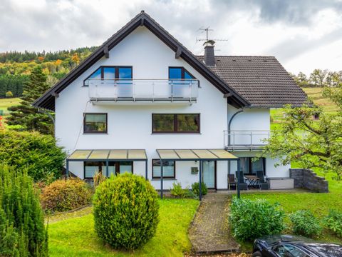Une maison moderne avec un grand jardin. Elle a des balcons et est entourée d'arbres.