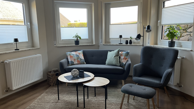 Un salon confortable avec un canapé gris et un fauteuil. Il y a des fenêtres lumineuses, un tapis et des tables d'appoint modernes.