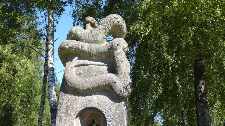 Eine hohe Skulptur aus Stein, die eine schlangenartige Form darstellt. Im Hintergrund sind grüne Bäume und ein blauer Himmel zu sehen.