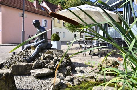 Statue en bronze d'un homme assis sur des pierres, entouré de roseaux et d'un petit étang, avec des maisons et un parasol en arrière-plan.