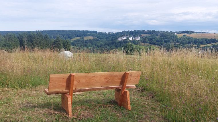 Eine Holzbank steht auf einer Wiese mit Blick auf eine hügelige Landschaft und ein Schloss in der Ferne.