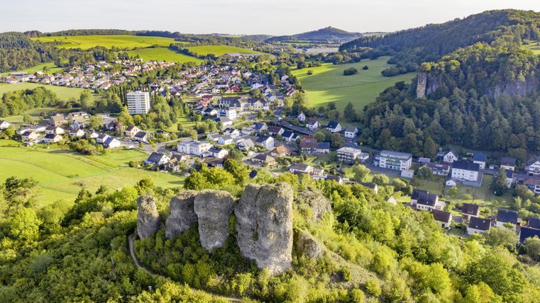 Blick von oben auf die Stadt Gerolstein mit zahlreichen Gebäuden und der Felsformation "Auberg", bestehend aus 5 nebeneinander aufragenden Felsen, im Vordergrund. Die Stadt ist Bergen und Bäumen umgeben.