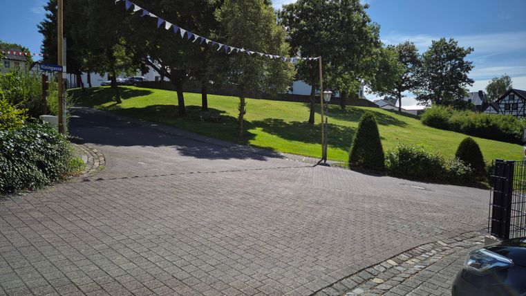 Eine gepflasterte Straße mit einer grünen Wiese und einigen Bäumen im Hintergrund. Im Himmel ist ein klarer blauer Himmel zu sehen.
