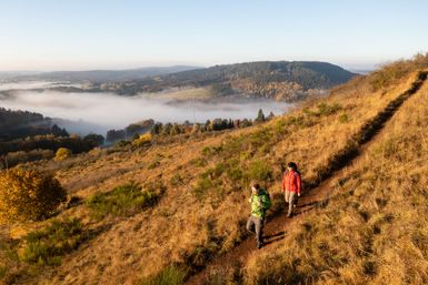Zwei personen wandern auf einem schmalen Wanderweg, dahinter Herbstlandschaft
