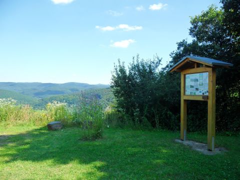 Une belle vue sur des collines vertes et un ciel bleu. Au premier plan se trouve un panneau d'information entouré d'herbe et de buissons.