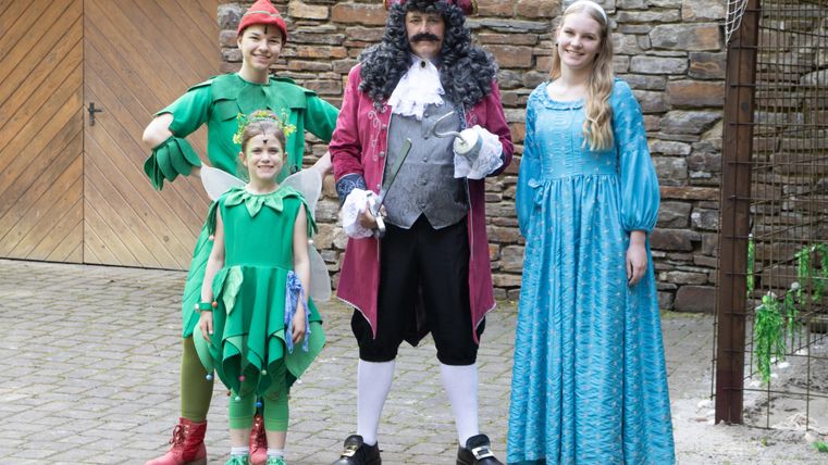 Eine Gruppe von Personen in bunten Kostümen steht vor einer Steinmauer. Sie stellen Charaktere aus einem Märchen dar, darunter ein Junge in einem grünen Kostüm und ein Kapitän in einem roten Cape.