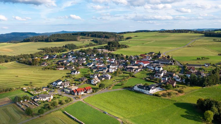 Eine malerische Landschaft mit einem kleinen Dorf umgeben von grünen Feldern. Sanfte Hügel und blauem Himmel schaffen eine ruhige Atmosphäre.