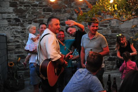 A cheerful group of people is celebrating outdoors while a musician plays with a guitar. String lights create a cozy atmosphere.
