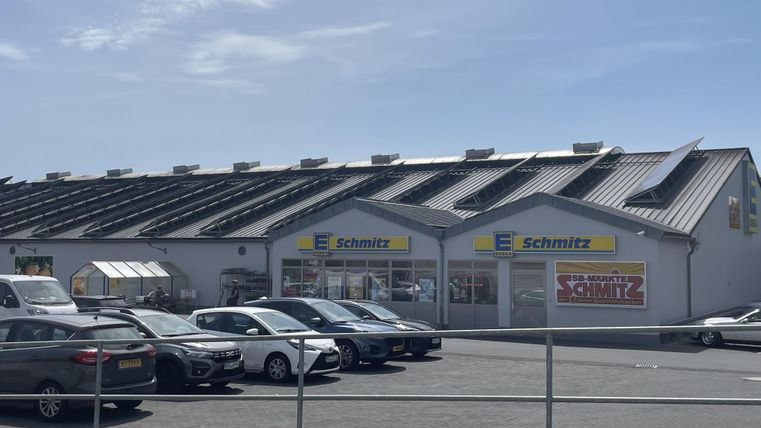 Un bâtiment commercial nommé "Schmitz" avec un toit moderne. Devant le bâtiment, plusieurs voitures sont garées sur un grand parking.