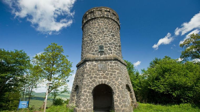 Een historische stenen toren staat omringd door weelderig groen en een blauwe lucht. De zon schijnt, en de wolken zijn gedeeltelijk zichtbaar.