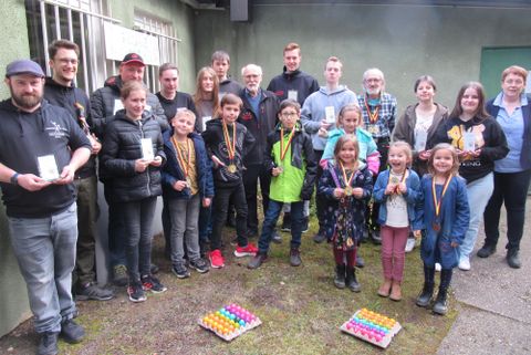 Eine Gruppe von Kindern und Erwachsenen steht draußen und hält Auszeichnungen in den Händen. Im Vordergrund sind bunte Eier gestapelt.
