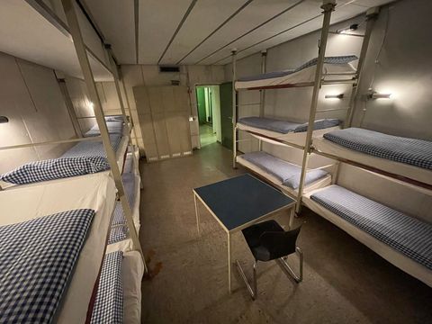 Ein einfacher Schlafsaal mit Etagenbetten und einem Tisch in der Mitte. Die Wände sind hell und es gibt einige Lichter an den Betten.