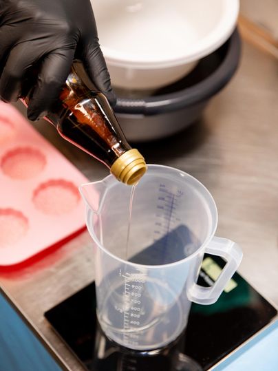 A person wearing black gloves is pouring a liquid from a bottle into a measuring cup. In the background, more kitchen utensils can be seen.